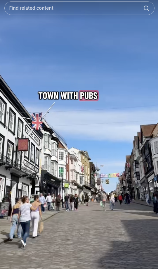 A busy UK high street lined with pubs and shops under blue skies, with people walking along the cobblestone road.